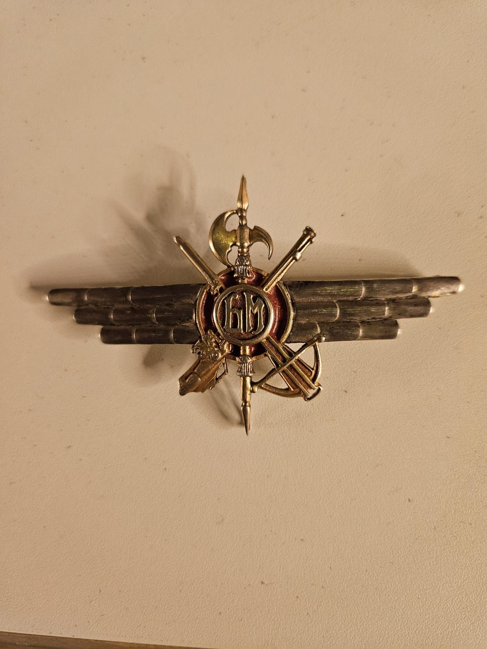 Emblema de latón y plata: alas, alabarda, ballesta y arcabuz cruzados.